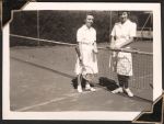 Long shot of two women standing on a tennis court, one of either side of the net. They wear white tennis attire and both face the camera. One of the women holds her tennis racket against the top of the net, the other holds it at her side. Women's Hospital, Circa 1946