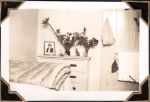 Medium shot of an indoor room with a bed, blanket, a door with a towel hung on the back and drawers with two photo frames on top, two vases with flowers and some decorative figures. Night Duty Room, Circa 1946