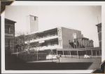 Long shot of three buildings. One is featured in the centre and two are on either side. There is a sheltered walkway with some people dressed in white sitting underneath it. In the foreground there are two tennis courts with nets next to a hedge and a tree. Women's Hospital, Circa 1946