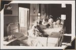 Long shot of a patient room. A patient can be seen in one of the beds with two staff members and some visitors sitting around them. There are two people in chairs close to the camera, the bed is behind them and the remaining people are on the other side of the bed. Women's Hospital, Circa 1946