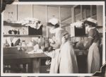 Medium shot of two staff members in uniform washing and changing two infant patients. They both wear masks over their mouth and nose and stand at a bench with sinks, cabinets and towels. Women's Hospital, Circa 1946