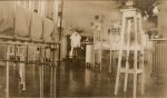 Long shot of a Nurse standing in the background of a ward with cots on both sides, a pot plant stand with two plants in the foreground. Ward Scene, 1928