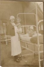 Long shot of a Nurse in a ward standing holding an infant alongside a young patient in a bed posing for the camera. Ward Scene, 1928