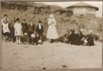 Long shot of a Nurse standing with a group of young patients, and a group of adults sitting on a beach, a small cliff and wooden rotunda in the background. At the Beach, 1928