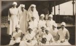 A long shot of a large group of Nurses standing and sitting outside with a young patient sitting, held by a Nurse, all posing for the camera. Staff Portrait, 1928