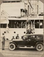 A long shot of a navy ship with adults walking down the gangplank, a car in the foreground on a wharf. There are groups of adults on the boat and wharf. Dockyard, 1928