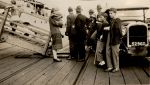 A long shot of adults and children standing on a wharf near the gangplank with a car in the foreground and a ship in the background. Dockyard, 1928