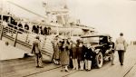 A long shot of adults and children standing on a wharf near the gangplank with a car in the foreground and a navy ship in the background. Dockyard, 1928