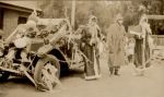 A long shot of three people in costume. Two are as Father Christmas. They are all standing beside a car that has been decorated with balloons and streamers. A small child can be seen in the background watching. Christmas Celebrations, 1928