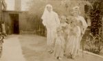 A long shot of a Nurse and four children standing outside in a garden dressed in costumes. One child is standing with a crutch. Dress Ups, 1928