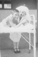 Medium shot of a Nurse standing next to a child lying in a cot, posing for the camera. Nurse and Child, Circa 1940