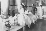 A long shot of a ward area with two staff members in uniform and an adult standing alongside five hospital beds with four children sitting in each. A child with their arm in a sling is standing beside a bed smiling for the camera. A table with a vase of flowers is left of camera. Elizabeth Ward, 1904