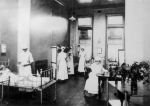 A long shot of a ward area three staff members in uniform standing alongside children sitting in three hospital cots. Another staff member in uniform is sitting writing notes on a table. A trolley with vases of flowers right of camera. Nurses in Ward, 1910