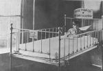 A mid shot of a child sitting in a hospital cot. Two signs are attached to both ends of the cot. Child in Cot, Circa 1900