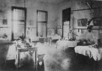 A long shot of a ward area three staff members in uniform standing alongside children laying or sitting in five hospital beds. A table left of camera has ornaments and a vase of flowers, two bench seats on either side of the table. Nurses in Ward, Circa 1890