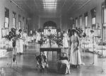 A long shot of a ward area with approximately ten hospital cots with children either laying down or sitting right and left of camera. Seven staff members in uniform are standing alongside the cots. Two children sitting in chairs in front of a table with vases of flowers are in foreground. Snowball Ward, 1908