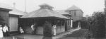 A long shot taken outside of a pavilion, two adults wearing white coats two staff members in uniform all standing two left of camera and two front of pavilion. Another staff member in uniform is sitting holding an infant, one of the adults and a staff member in uniform are holding children in their arms. A building in the background, tree in the foreground and trees right of camera. Isolation Pavilion, Circa 1920