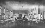 A long shot of a ward area, hospital cots are right and left of camera. Three children are sitting on left and right of a table with vases of flowers. Five staff members in uniform and an adult wearing a suit are standing directly behind the seated children, two staff members are holding infants in their arms. Francis Ormond Ward, Circa 1908