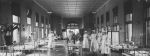 A long shot of the Snowball ward with hospital cots right and left of camera children are laying on each cot, eight staff members in uniform standing mostly in centre of ward, a table with vases of flowers in the foreground. Snowball Ward, 1913