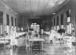 A long shot of a ward area, hospital cots are right and left of camera. Children are sitting in the hospital cots looking at the camera. Two staff members in uniform are standing right and left of camera alongside the cots. A table with a vase of flowers is centre of ward, three stands containing vases of flowers or pot plants are left and right of camera. Ward Area, Circa 1923