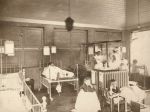 A long shot of the Isolation ward three children are sitting in hospital cots, one child is sitting in a chair, another child right of camera is standing alongside a cot. Two staff members in uniform are standing looking right of camera. Isolation Ward, 1908