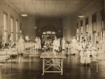 A long shot of the Snowball ward with twelve hospital cots children are sitting in the cots looking at the camera, eight staff members in uniform are standing alongside the cots, two are holding infants in their arms. Tables with vases of flowers in centre of ward. Snowball Ward, 1913