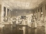 A long shot of a ward area, hospital cots are right and left of camera. Children are sitting in the hospital cots looking at the camera. Seven staff members in uniform are standing right and left of camera alongside the cots one is holding a child in their arms. Another staff member in uniform is sitting behind a desk with a child in their arms centre of ward, pot plants on stands are left and right of camera. Lower Surgical Ward, Circa 1920
