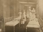 A long shot of a balcony area with children laying in six hospital cots, one child is sitting on a chair, three staff members in uniform are standing attending to the children. Balcony Ward Area, circa 1920
