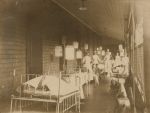 A long shot of a balcony area with children laying in hospital cots, five children are sitting on chairs, five staff members in uniform are standing looking at the camera. Sumner Balcony, Circa 1913