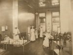 A long shot of a ward area three staff members in uniform standing alongside children sitting in three hospital cots. Another staff member in uniform is sitting writing notes on a table. A trolley with vases of flowers right of camera. Nurses in Ward, 1910