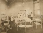 A long shot of a theatre three staff members in uniform and two adults wearing white coats are standing in the background one of the adult is leaning on a glass cabinet filled with medicine bottles. Surgical trolley and tables with medical equipment in the foreground. A sink left of camera with bowls on stands in front. Surgical Theatre, Circa 1919