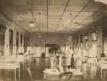 A long shot of the summer ward with hospital cots right and left of camera children laying on each cot, six staff members in uniform are standing alongside the cots right of camera, one child wearing a hospital gown is standing alongside a staff member, in the centre of the ward is a steam heater with a marble top with vases of flowers. Sumner Ward, 1901