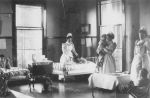 A long shot of a ward area with three staff members in uniform standing, one holding an infant, one attending a child in a hospital bed, the other staff member right of camera is alongside a child sitting on a chair. Sumner Ward, 1894