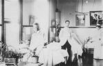 A long shot of a ward area with two staff members in uniform and an adult wearing a white coat standing in front of three hospital beds with three children laying in each. A table with potted plants is left of camera. Elizabeth Ward, 1894